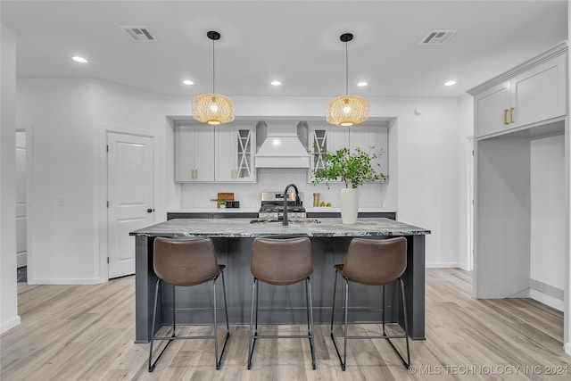 kitchen with light stone countertops, premium range hood, a kitchen island with sink, sink, and decorative light fixtures