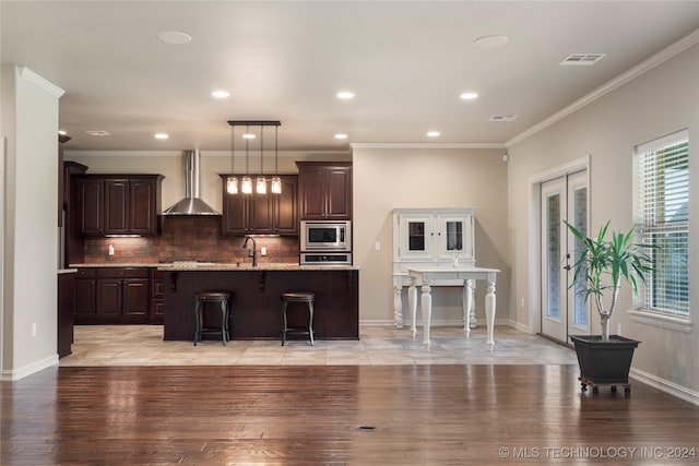 kitchen with a kitchen breakfast bar, wall chimney exhaust hood, stainless steel appliances, light hardwood / wood-style flooring, and an island with sink