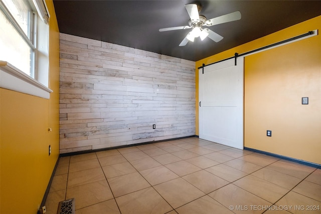 unfurnished bedroom with a barn door, ceiling fan, light tile patterned floors, and wood walls