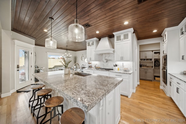 kitchen with custom range hood, sink, pendant lighting, a center island with sink, and white cabinets