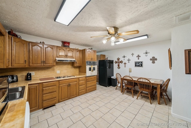 kitchen featuring brown cabinets, oven, freestanding refrigerator, light countertops, and under cabinet range hood