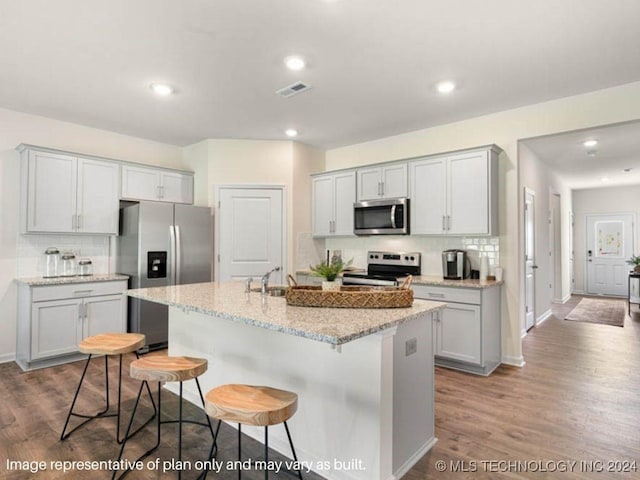 kitchen with light stone countertops, decorative backsplash, a center island with sink, appliances with stainless steel finishes, and light wood-type flooring