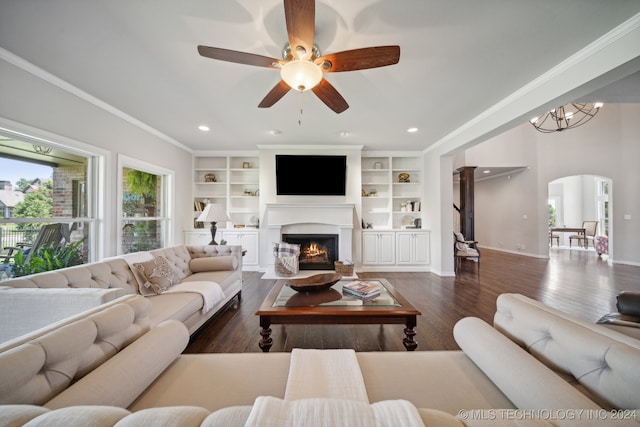 living room with dark wood-type flooring, ornamental molding, ceiling fan with notable chandelier, and built in shelves