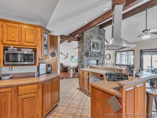 kitchen with a stone fireplace, vaulted ceiling with beams, island exhaust hood, a textured ceiling, and appliances with stainless steel finishes