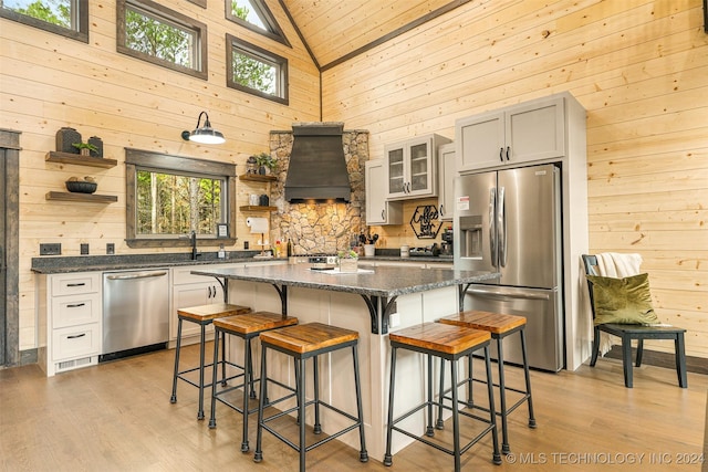 kitchen featuring custom exhaust hood, a breakfast bar, stainless steel appliances, wooden walls, and high vaulted ceiling