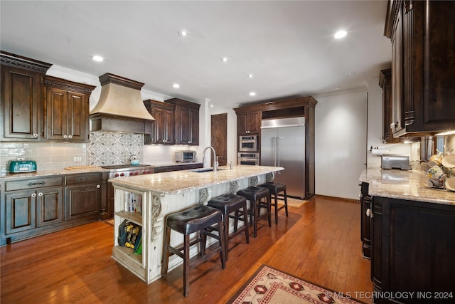 kitchen with dark wood-type flooring, a kitchen bar, a kitchen island with sink, custom range hood, and appliances with stainless steel finishes