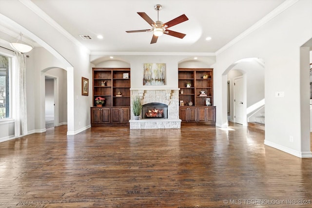 unfurnished living room with dark wood-type flooring, a stone fireplace, ceiling fan, built in shelves, and ornamental molding