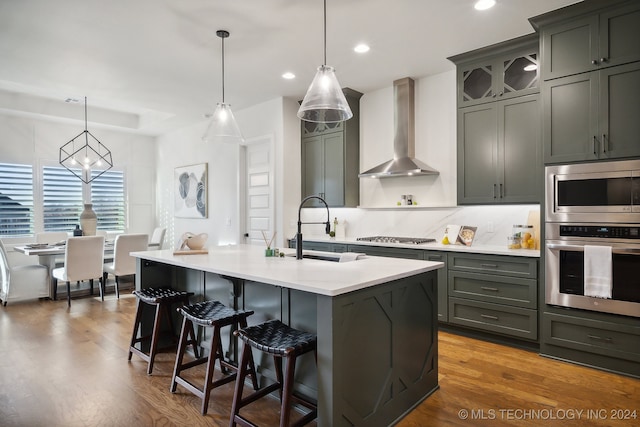 kitchen with pendant lighting, a kitchen island with sink, wall chimney range hood, sink, and appliances with stainless steel finishes