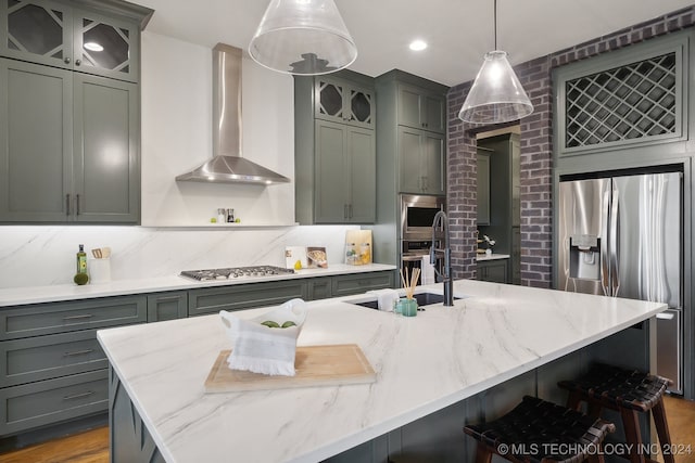 kitchen with pendant lighting, wall chimney range hood, stainless steel appliances, and a kitchen island with sink