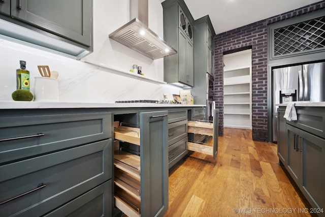 kitchen featuring light hardwood / wood-style floors, brick wall, wall chimney range hood, stainless steel fridge with ice dispenser, and gray cabinets