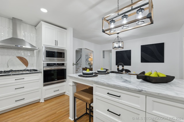 kitchen featuring white cabinetry, stainless steel appliances, wall chimney range hood, decorative backsplash, and light wood-type flooring