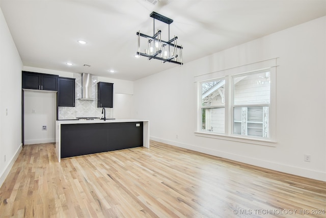 kitchen with tasteful backsplash, a kitchen island with sink, wall chimney range hood, decorative light fixtures, and light hardwood / wood-style floors