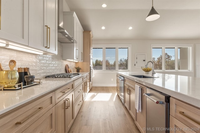 kitchen featuring wall chimney range hood, sink, light brown cabinetry, appliances with stainless steel finishes, and decorative light fixtures