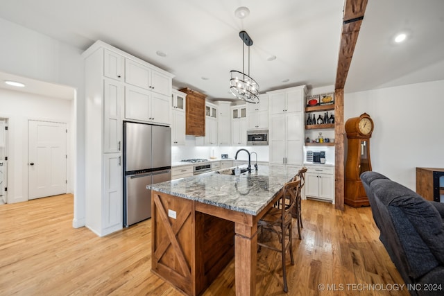 kitchen with light stone countertops, decorative light fixtures, a center island with sink, appliances with stainless steel finishes, and light wood-type flooring