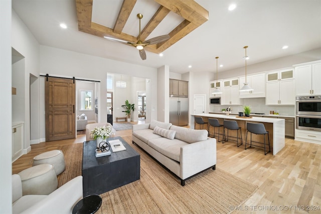 living room featuring a barn door, ceiling fan, a high ceiling, light hardwood / wood-style flooring, and beamed ceiling