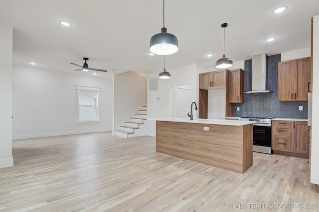 kitchen featuring wall chimney exhaust hood, stainless steel range, pendant lighting, a center island with sink, and light hardwood / wood-style floors