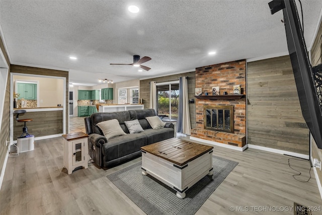 living room with ceiling fan, light hardwood / wood-style floors, a textured ceiling, a brick fireplace, and wood walls