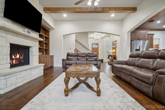 living room with dark hardwood / wood-style flooring, ceiling fan with notable chandelier, built in features, beamed ceiling, and a stone fireplace