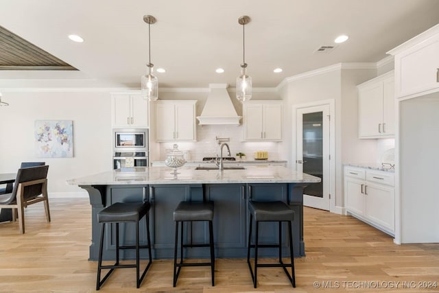 kitchen with custom range hood, stainless steel appliances, a kitchen island with sink, decorative light fixtures, and white cabinets