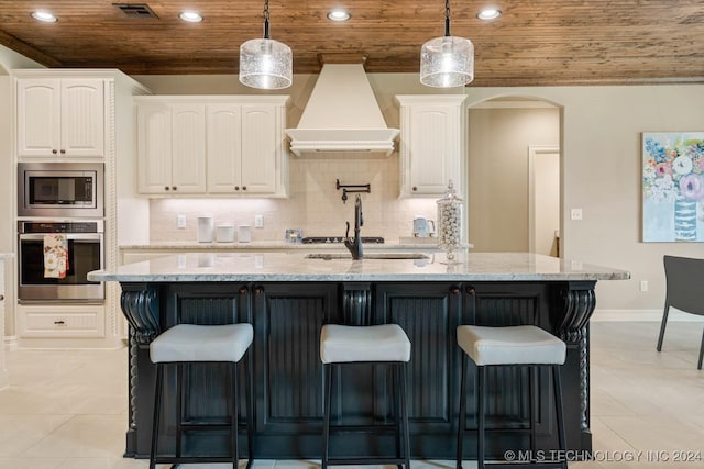 kitchen featuring a kitchen island with sink, premium range hood, appliances with stainless steel finishes, white cabinetry, and wood ceiling