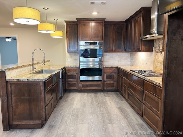 kitchen with kitchen peninsula, stainless steel appliances, wall chimney range hood, pendant lighting, and light hardwood / wood-style flooring