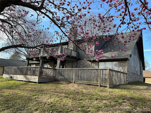 back of property with a balcony, roof with shingles, a yard, stone siding, and a deck