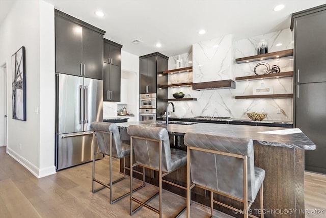 kitchen with a center island with sink, dark countertops, stainless steel appliances, a kitchen bar, and open shelves