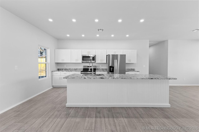 kitchen with white cabinetry, light stone counters, light wood-type flooring, appliances with stainless steel finishes, and an island with sink