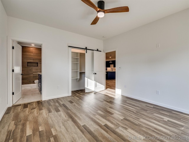 unfurnished bedroom with ensuite bathroom, ceiling fan, a barn door, light hardwood / wood-style floors, and a closet