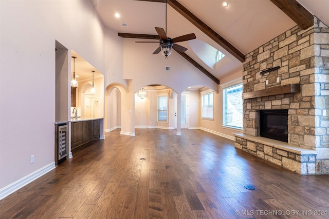 unfurnished living room featuring wine cooler, dark wood-type flooring, a stone fireplace, ceiling fan, and beam ceiling