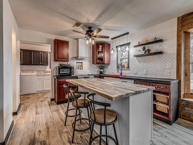 kitchen with ceiling fan, wall chimney range hood, butcher block countertops, washer / clothes dryer, and a kitchen island