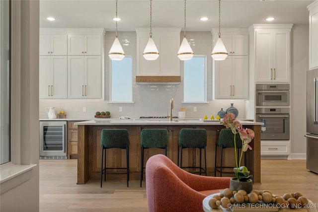 kitchen with tasteful backsplash, beverage cooler, a kitchen island with sink, pendant lighting, and white cabinets