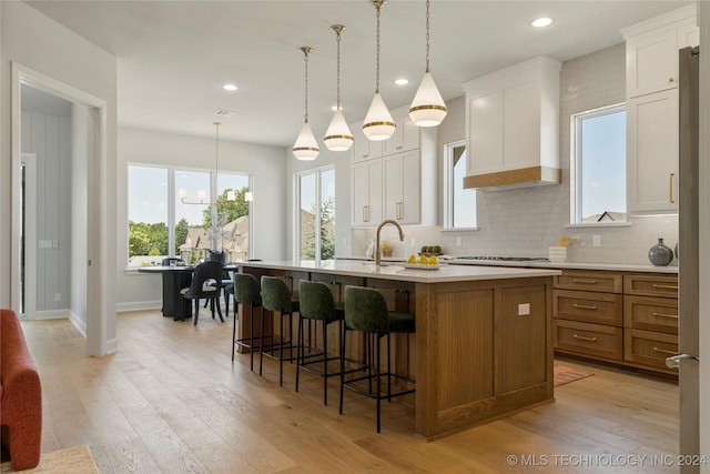 kitchen featuring custom exhaust hood, hanging light fixtures, light hardwood / wood-style flooring, an island with sink, and white cabinetry