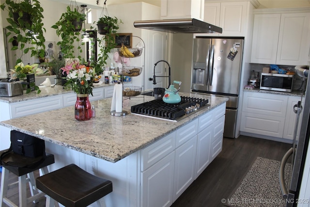 kitchen with white cabinetry, stainless steel appliances, a kitchen island with sink, wall chimney exhaust hood, and light stone counters