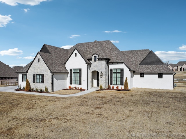 french country inspired facade with a front yard, stone siding, and roof with shingles