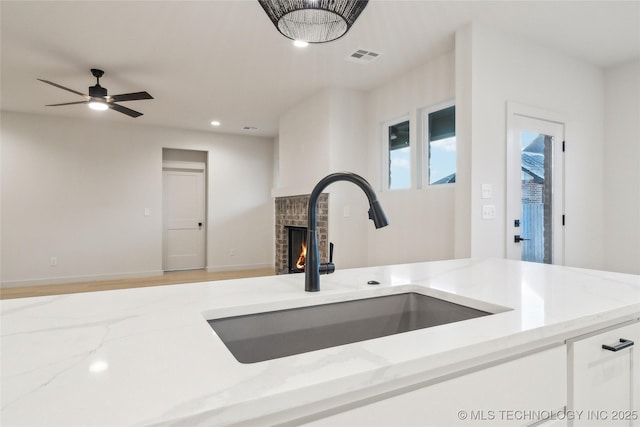 kitchen featuring white cabinetry, sink, ceiling fan, light stone countertops, and a brick fireplace