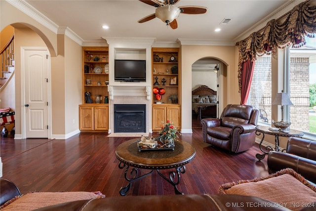 living room featuring ceiling fan, dark wood-type flooring, built in features, a tiled fireplace, and ornamental molding