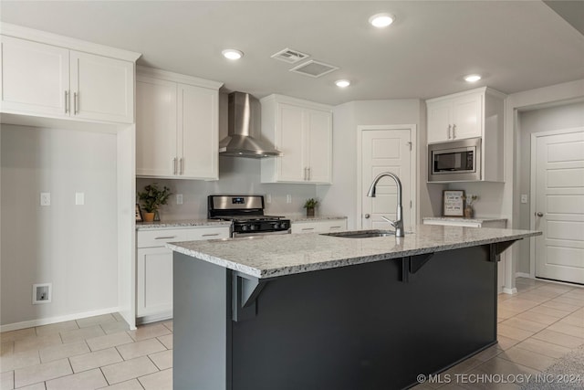 kitchen with white cabinetry, wall chimney range hood, a center island with sink, light tile patterned flooring, and appliances with stainless steel finishes