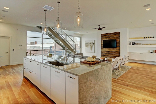 kitchen featuring a kitchen island with sink, hanging light fixtures, light hardwood / wood-style flooring, light stone countertops, and white cabinetry