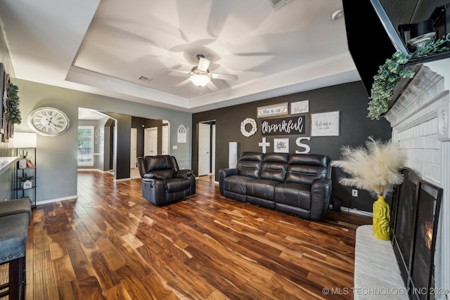 living room featuring a raised ceiling, ceiling fan, dark wood-type flooring, crown molding, and a fireplace