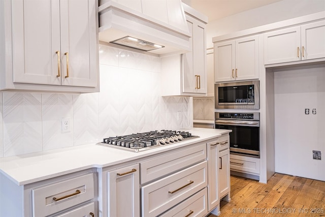 kitchen with light wood-style flooring, appliances with stainless steel finishes, light countertops, premium range hood, and backsplash