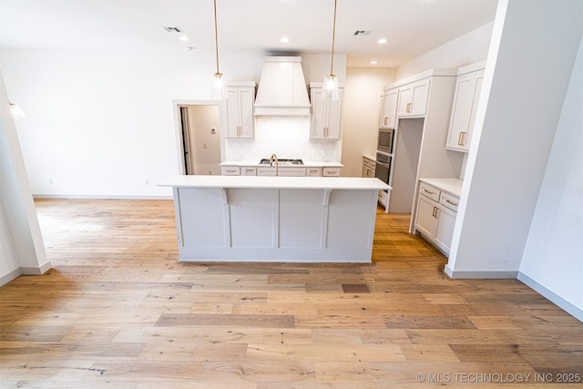 kitchen with custom exhaust hood, light wood finished floors, backsplash, a kitchen island with sink, and built in microwave