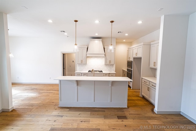 kitchen with premium range hood, appliances with stainless steel finishes, backsplash, light wood finished floors, and a center island with sink