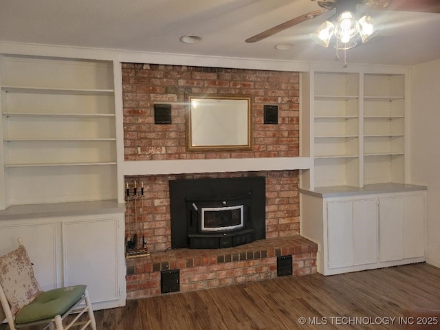 unfurnished living room with a wood stove, ceiling fan, built in shelves, and dark hardwood / wood-style flooring