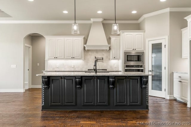 kitchen with custom exhaust hood, a kitchen island with sink, white cabinets, hanging light fixtures, and appliances with stainless steel finishes