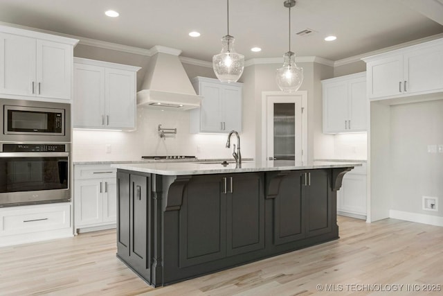 kitchen with custom exhaust hood, stainless steel appliances, a kitchen island with sink, decorative light fixtures, and white cabinets