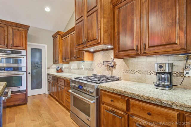 kitchen with appliances with stainless steel finishes, decorative backsplash, light stone counters, custom range hood, and light hardwood / wood-style flooring