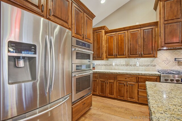 kitchen featuring vaulted ceiling, appliances with stainless steel finishes, backsplash, light stone counters, and light wood-type flooring