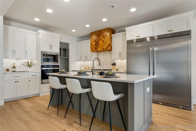 kitchen featuring backsplash, a kitchen island with sink, sink, built in appliances, and white cabinetry