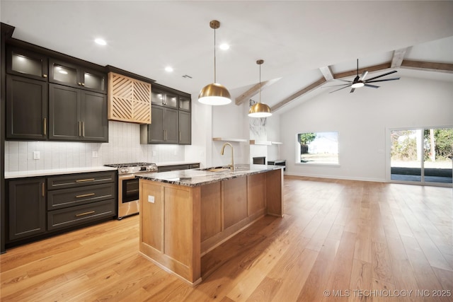 kitchen with light wood-type flooring, stainless steel gas range oven, dark stone counters, beam ceiling, and a center island with sink